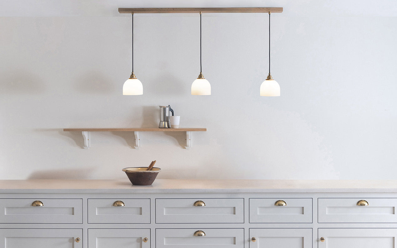 Glass Bue - Linear Trio Light Long with three frosted glass shades hanging from a long wooden bar above a white kitchen counter and wooden shelf