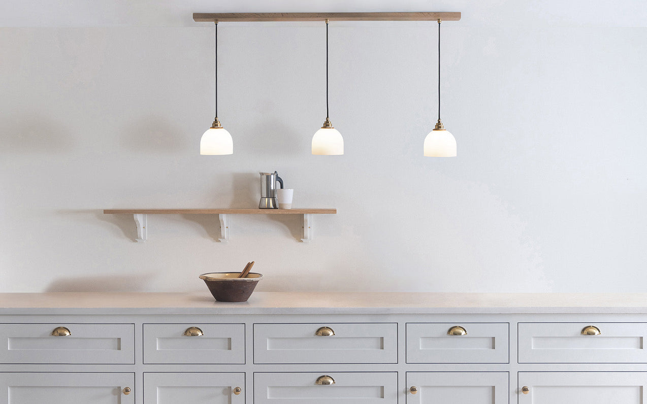 Glass Bue - Linear Trio Light Long with three frosted glass shades hanging from a long wooden bar above a white kitchen counter and wooden shelf